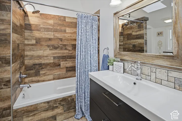White bathtub with chrome fixtures, wood-finish tile shower surround, contemporary vanity with integrated sink, patterned tile backsplash, and a large framed mirror