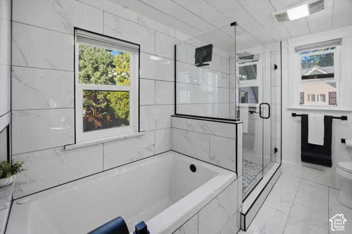 Freestanding soaking tub with white subway tile surround, glass-enclosed shower featuring a patterned tile floor, and a single window with a marble-finish sill