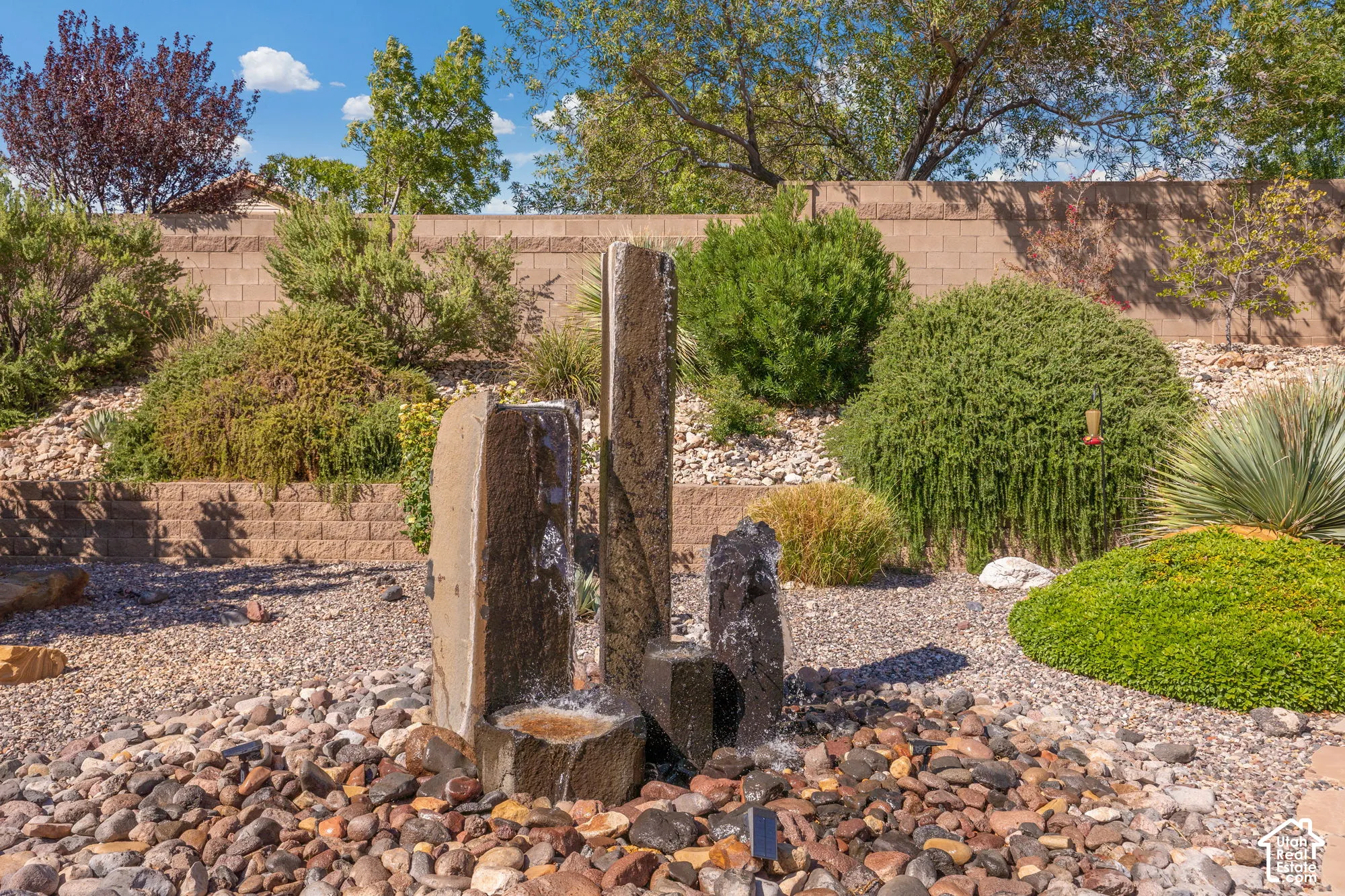 Stone water feature with varied stone and gravel ground cover, surrounded by tiered landscaping with diverse plantings, and a block privacy wall
