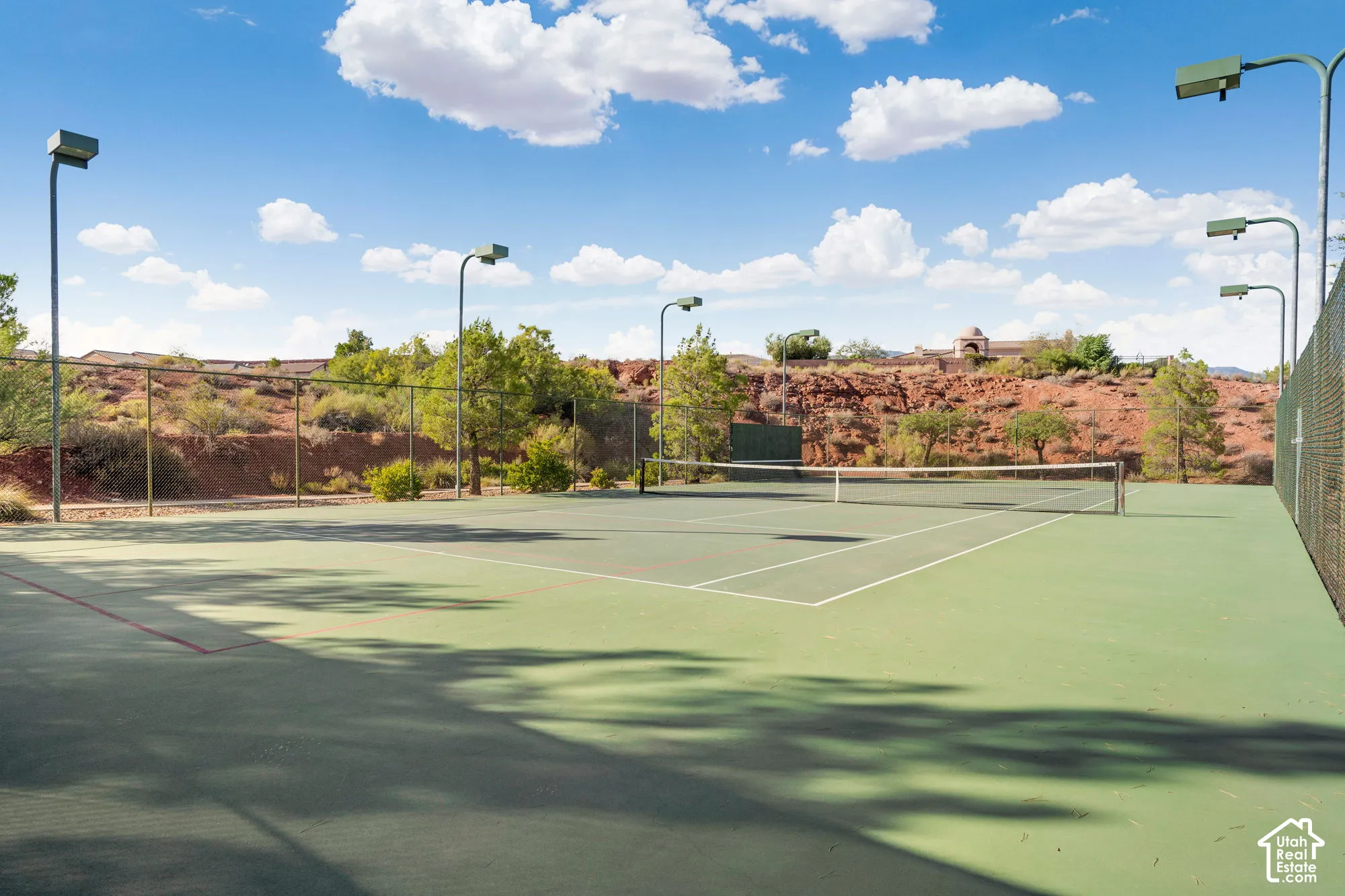Hard surface tennis court with painted lines, surrounded by chain-link fencing, featuring multiple overhead lighting fixtures