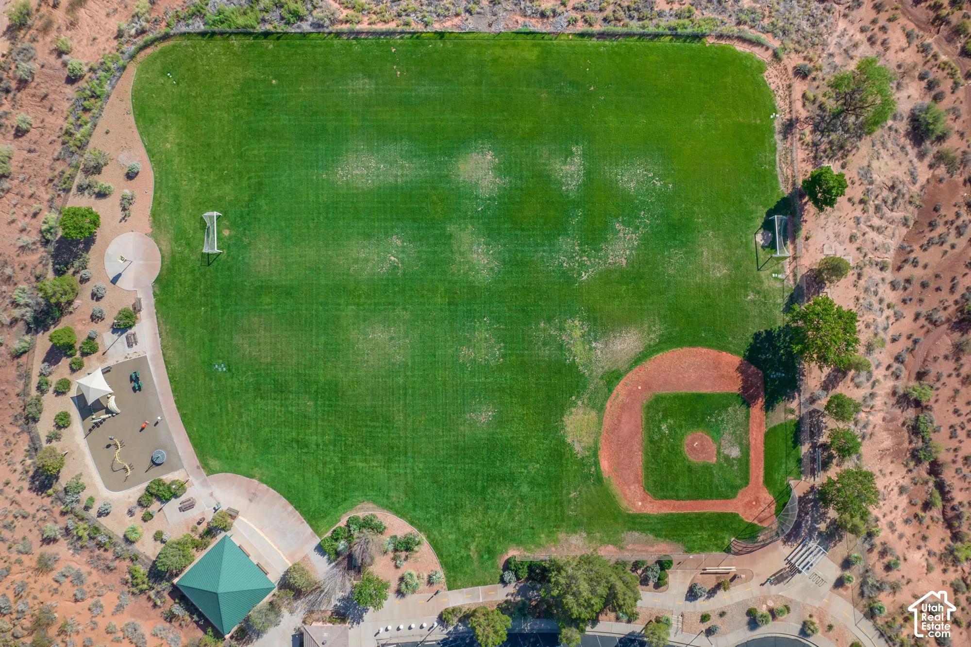 Expansive green space featuring a baseball diamond, two soccer goals, a shaded playground area, and a covered picnic structure