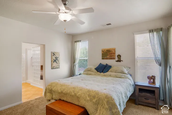 Carpeted bedroom featuring a ceiling fan with light, two windows with blinds, and an entryway to an adjoining room with glass block walls and tile flooring