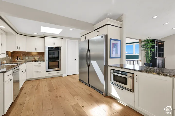Kitchen featuring wood-finish flooring, white cabinetry, integrated stainless steel appliances, a double wall oven, and a skylight