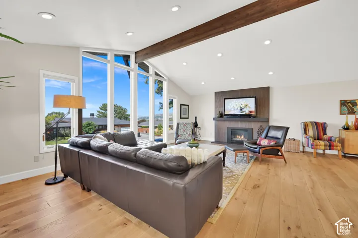 Vaulted ceiling with exposed wood beam, recessed lighting, expansive picture windows, LVP  flooring, and a fireplace with a wood-paneled surround