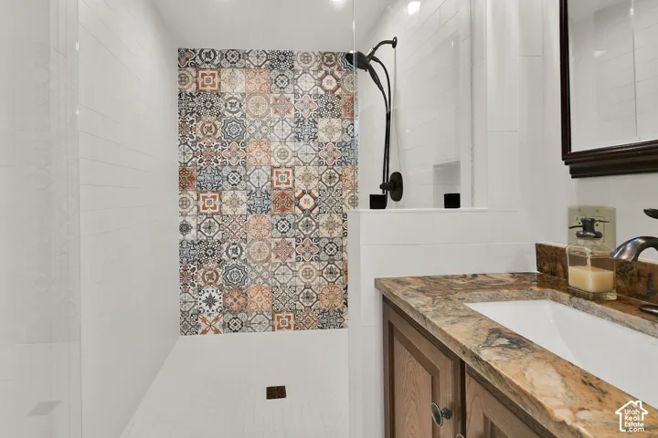 Walk-in shower with patterned wall tile, matte black shower fixtures, vanity with integral sink, wood-finish cabinetry, and stone countertop