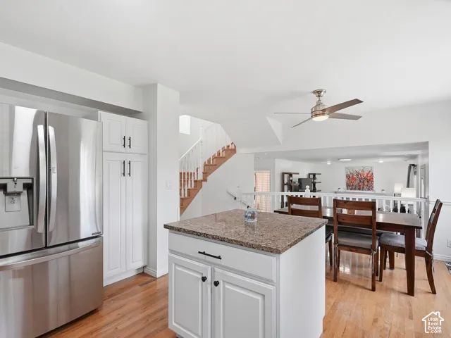 White kitchen cabinetry, stainless steel refrigerator, kitchen island with stone countertop, wood-finish flooring, and a ceiling fan