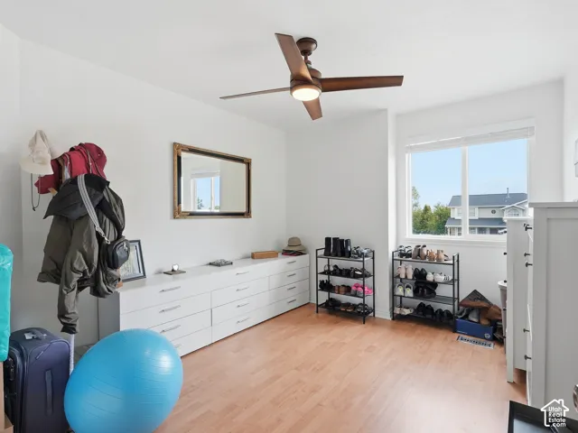 Bright secondary bedroom featuring warm wood-finish flooring, abundant natural light from dual windows, ceiling fan with integrated lighting, and extensive white cabinetry storage