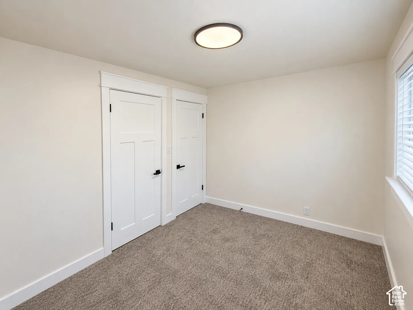 Neutral-toned walls, plush carpet flooring, white paneled doors, black door hardware, flush-mount ceiling light fixture, and a window with white blinds