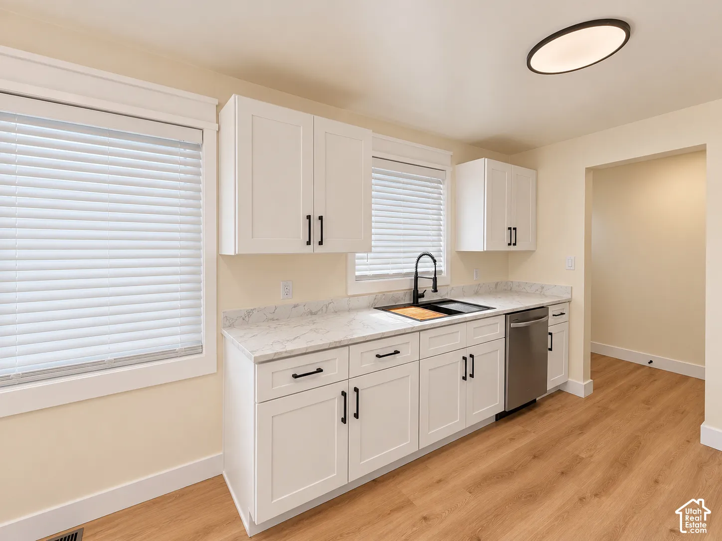 White shaker-style cabinetry with matte black hardware, light-toned countertops, stainless steel dishwasher, and wood-finish flooring