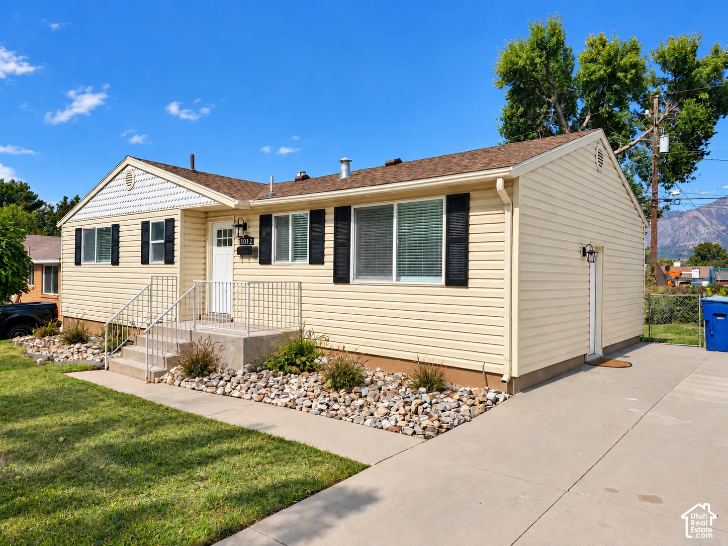 Cream-colored siding exterior with dark shutters, concrete walkway and driveway, front porch with white railings, and a peaked roofline