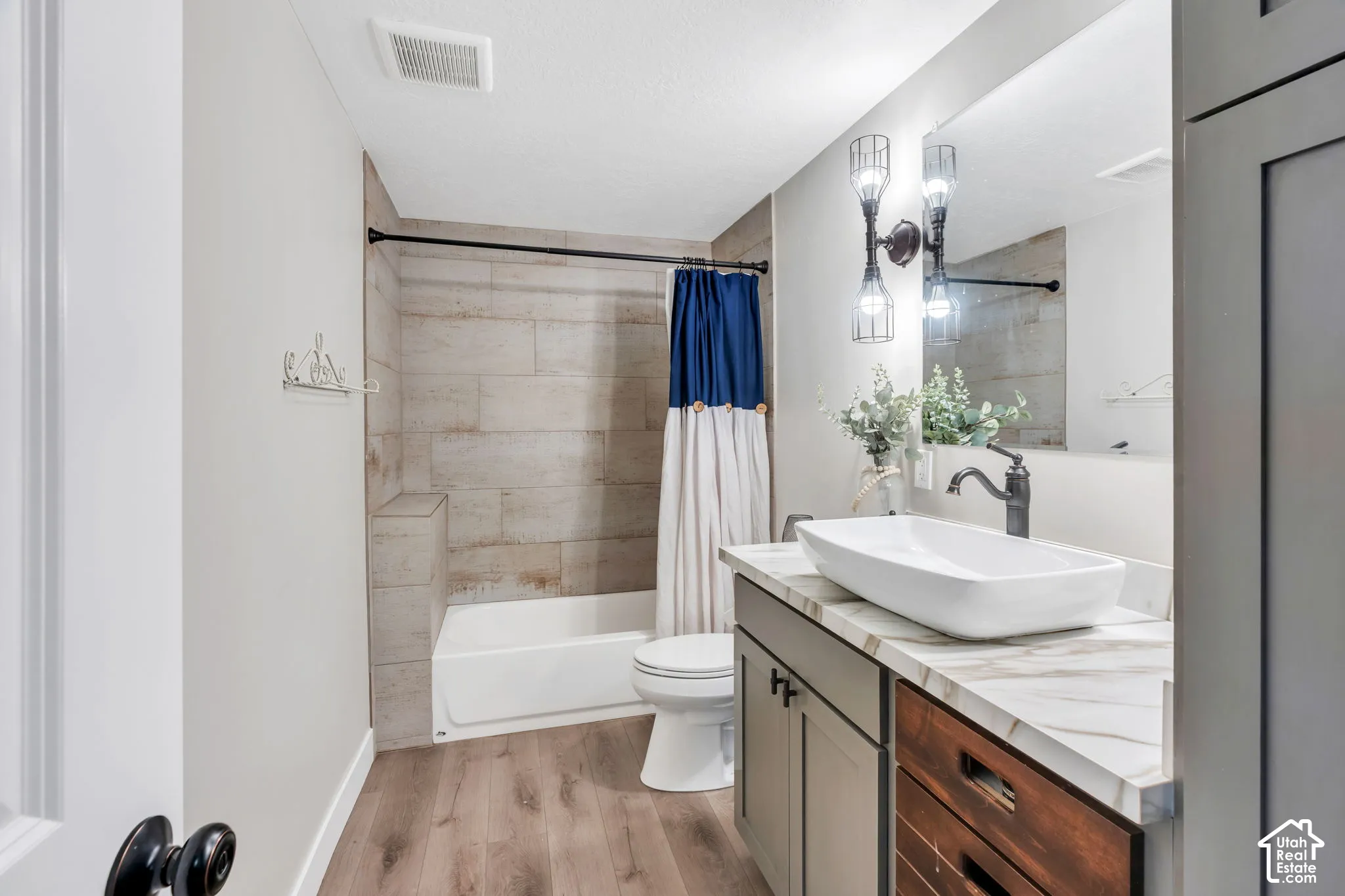 Wood-finish flooring, full bathtub with tiled surround, vessel sink, vanity with mixed-material cabinetry, and wall-mounted lighting