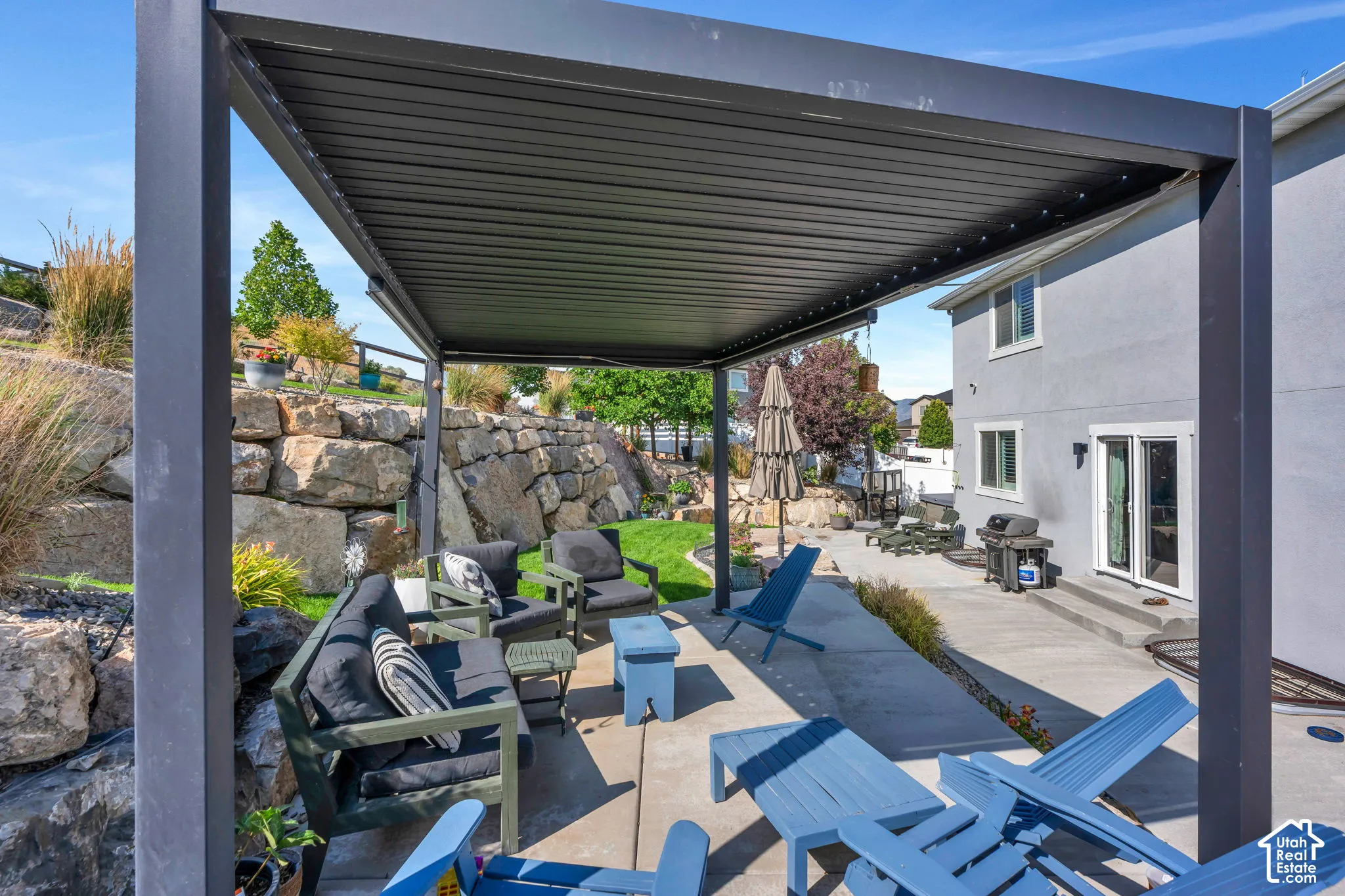 Paved patio featuring a louvered pergola, rock retaining wall, landscaped garden beds, and direct access to the interior via a sliding glass door