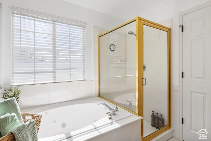 Primary Bathroom with White jetted soaking tub, glass-enclosed shower with gold-tone framing, white window blinds, and paneled interior door