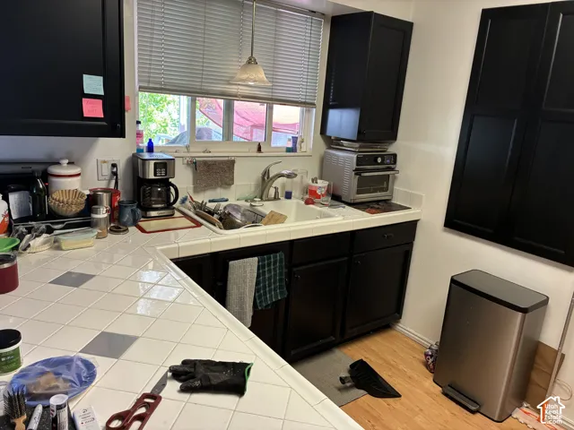 Kitchen featuring black cabinetry, light wood-finish flooring, a white tiled countertop with gray accents, a double-basin sink with a stainless steel faucet, and a window with horizontal blinds