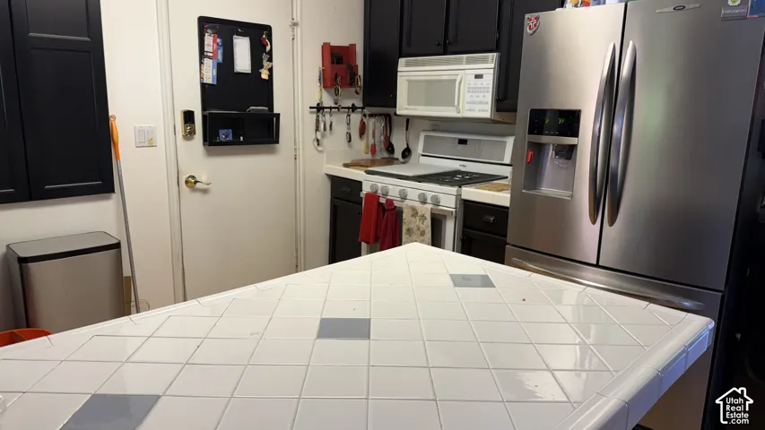 Kitchen featuring a tile countertop, white gas range, stainless steel refrigerator, white microwave, and dark cabinetry