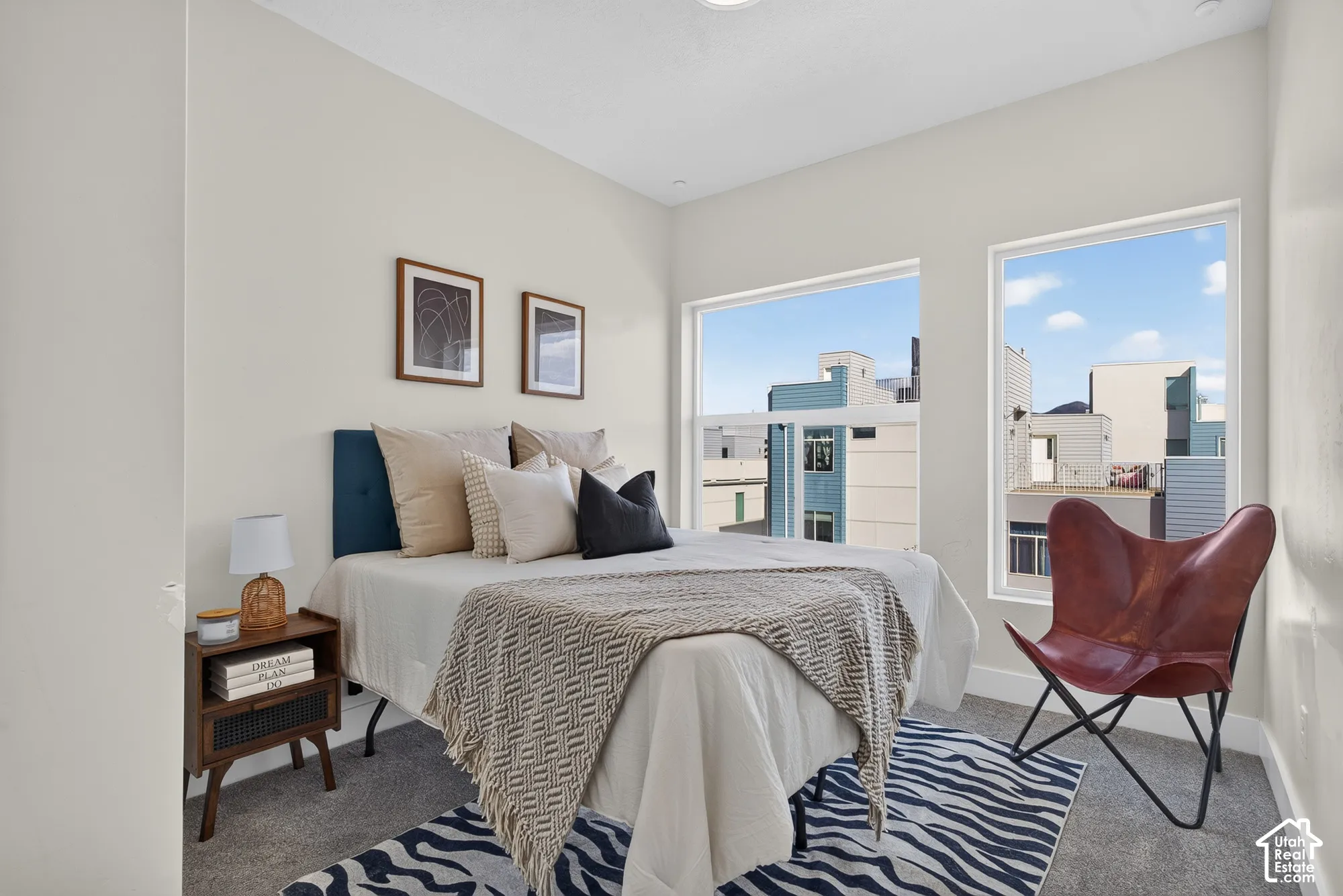 Light-toned wall finishes, carpet flooring, a dark wood-finish nightstand, and two large windows with white trim