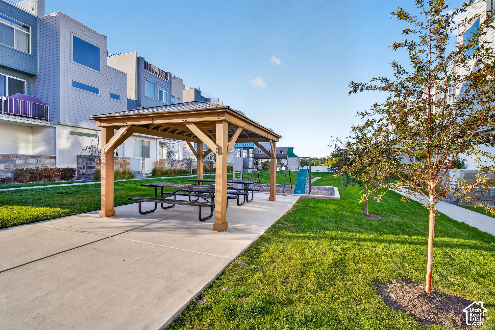 Wood-tone gazebo with picnic tables, adjacent to a manicured lawn, paved pathways, and a children's play area