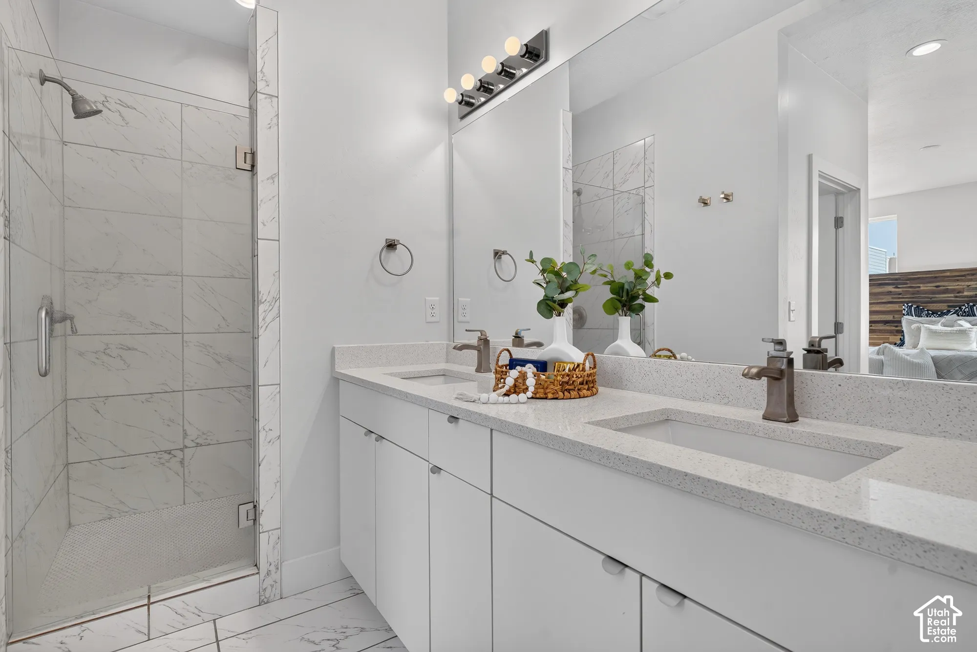 Tiled shower enclosure with a glass door, double vanity with integrated sinks, speckled countertops, and a wide mirror with overhead lighting