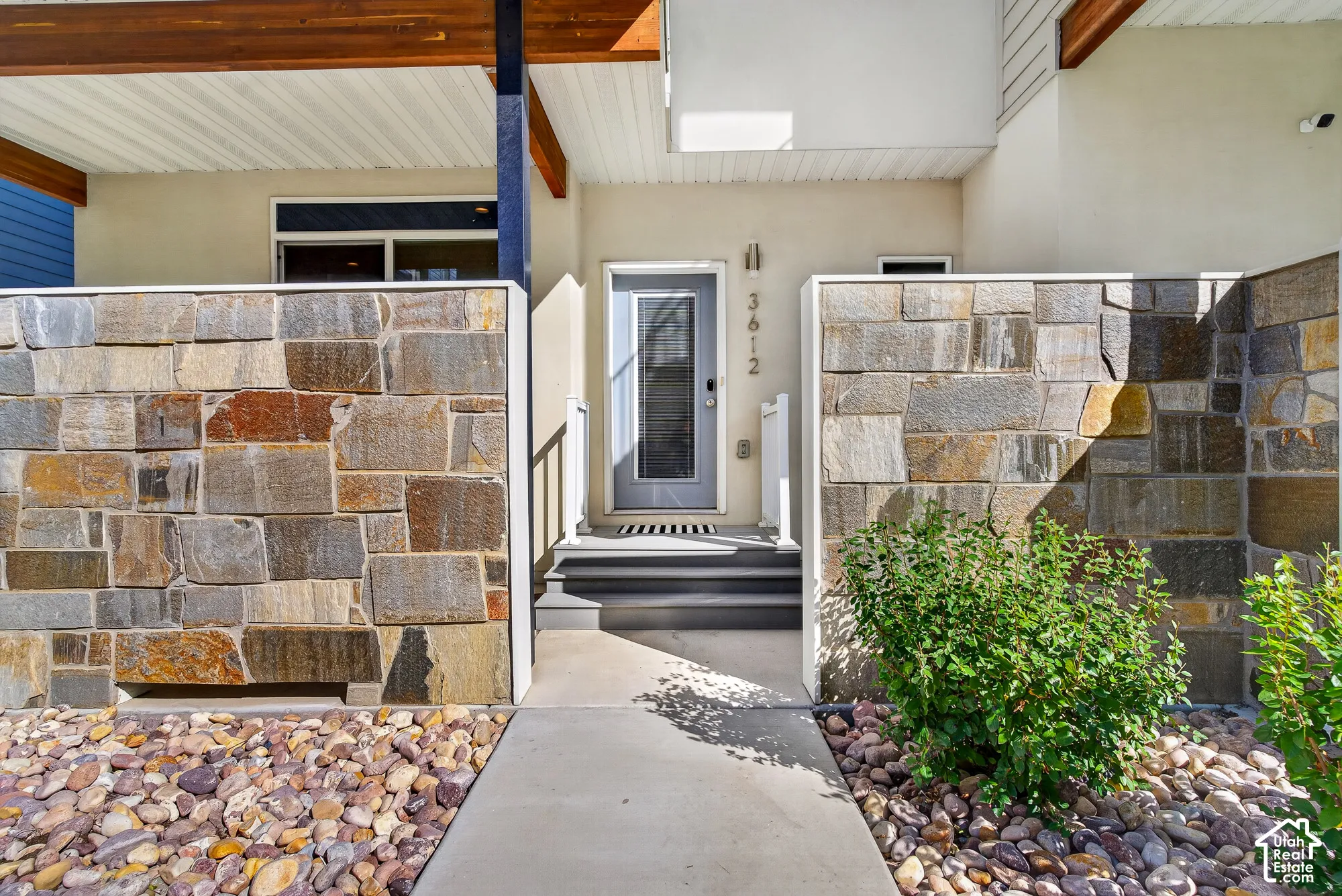 Stone facade accent walls, concrete walkway, multi-toned pebble landscaping, integrated outdoor lighting, and wood-beam architectural elements