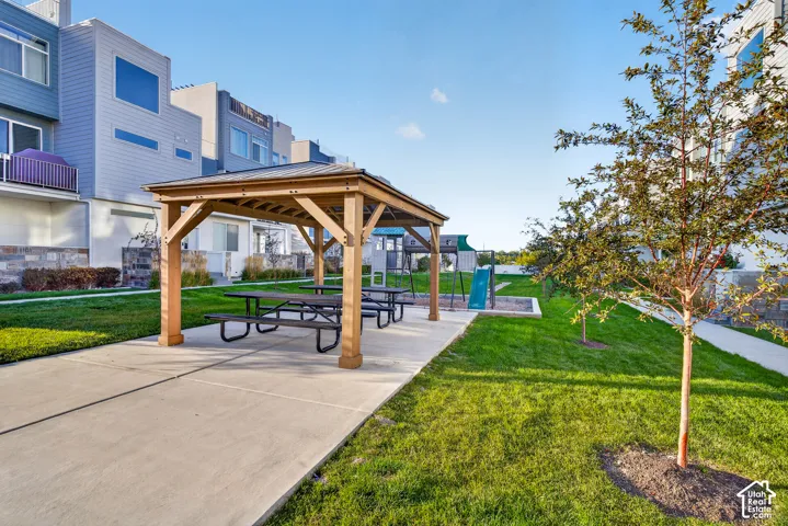 Wood-tone gazebo with picnic tables, adjacent to a manicured lawn, paved pathways, and a children's play area