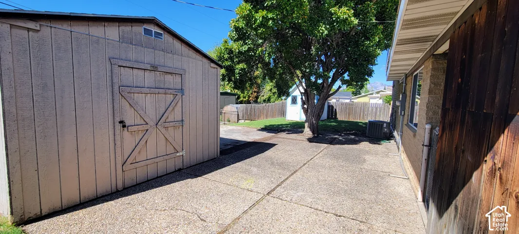 Storage shed with a barn-style door, concrete patio, mature trees, and a privacy fence