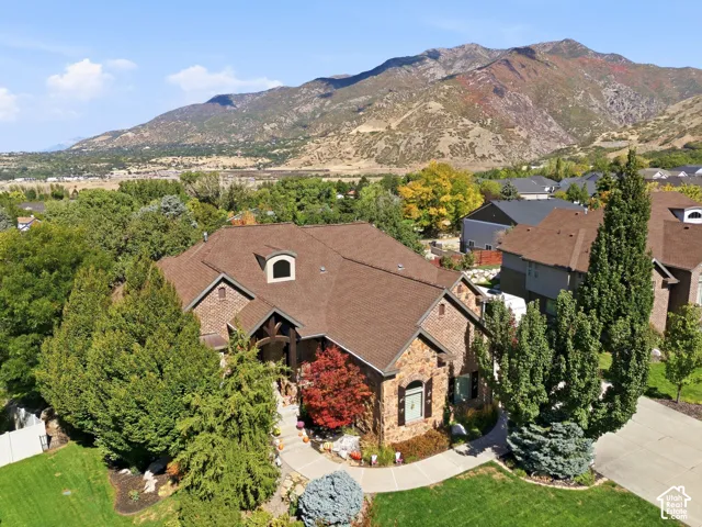 Stone facade residence featuring a brown shingle roof with dormer, mature landscaping, and a concrete driveway