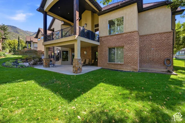 Two-story exterior with stucco and brick finishes, covered patio with stone-wrapped columns, upper-level balcony with metal railing, and a landscaped yard