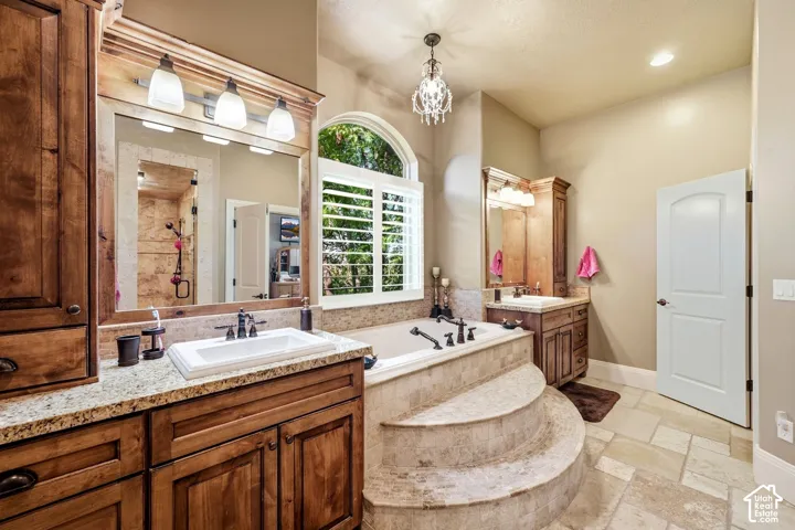 Soaking tub with tiled steps, dual vanities with granite countertops, wood cabinetry, and plantation shutters on an arched window