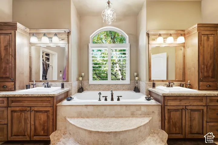 Dual wood-finish vanities with undermount sinks, a built-in soaking tub, a decorative chandelier, and an arched window with louvered shutters