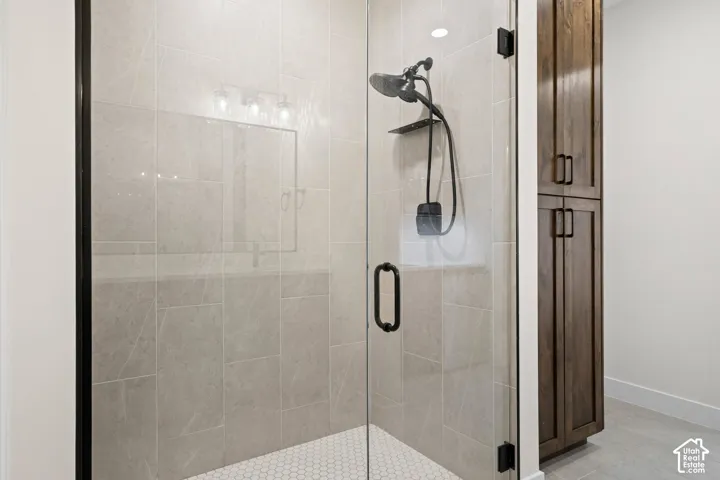 Glass-enclosed shower with a matte black shower system, light-toned wall tiling, white hexagonal floor tiling, and dark wood-finish cabinetry