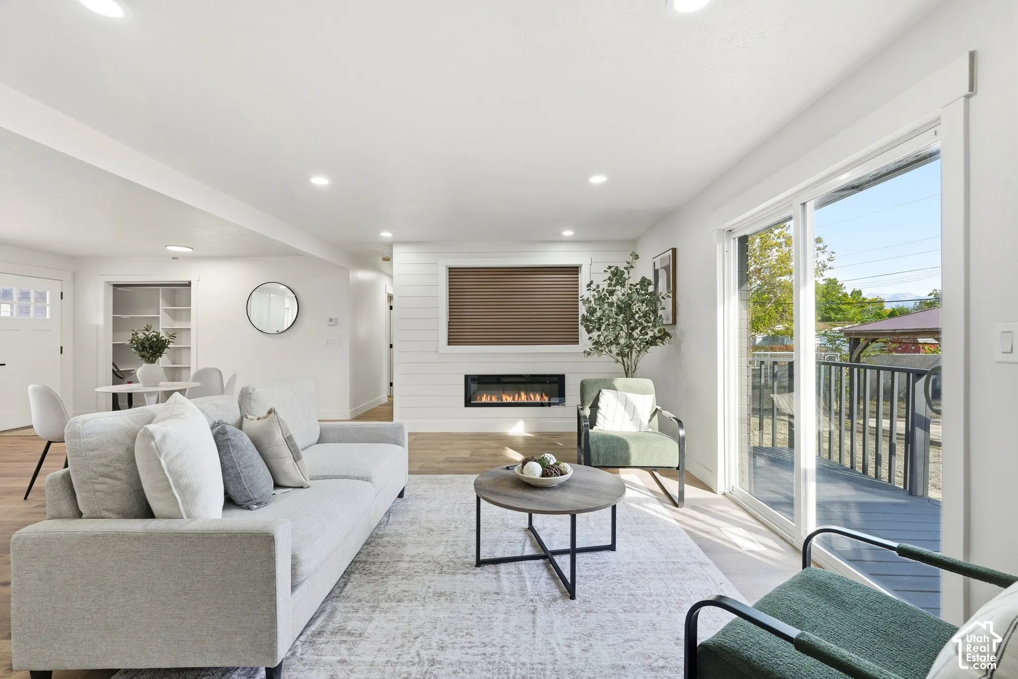 Open-concept living area featuring wood-finish flooring, a contemporary fireplace with shiplap surround, recessed lighting, and a sliding glass door opening to an exterior deck