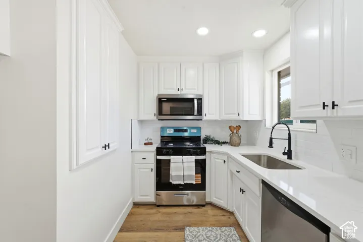 White cabinetry with black hardware, stainless steel appliances, kitchen sink with a black gooseneck faucet, light countertops, and wood-finish flooring