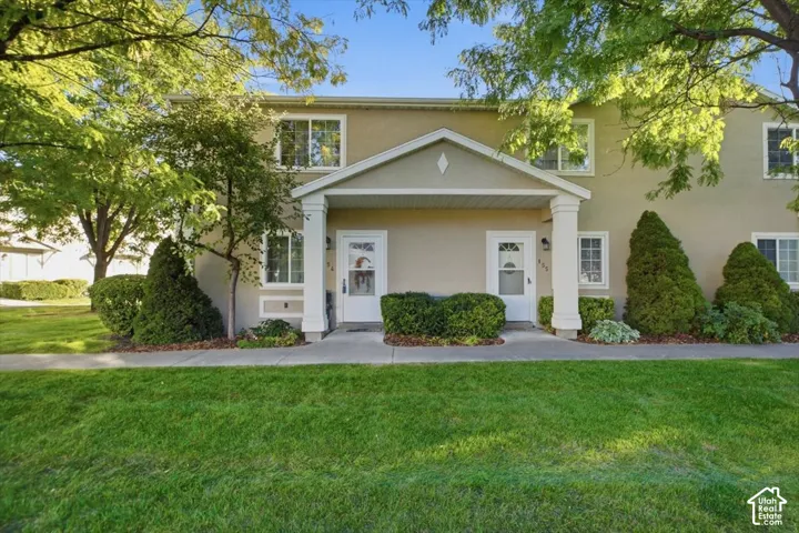 Two-story stucco exterior featuring white trim, a covered porch with columns, a well-maintained lawn, and mature landscaping