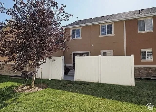 Manicured lawn with mature tree, white privacy fencing, two-story exterior with stucco finish, stone veneer foundation, and multiple windows