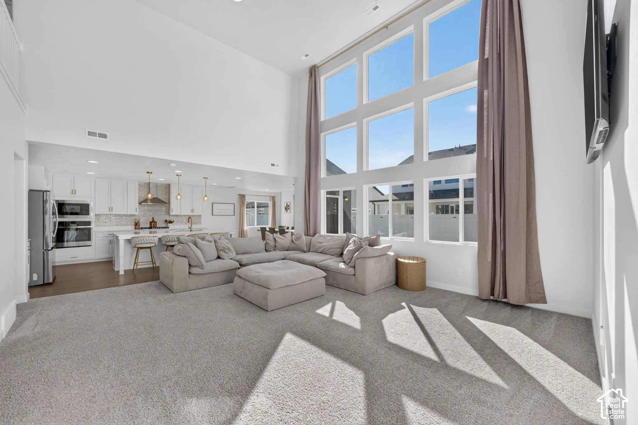Two-story volume ceiling, large multi-pane windows, light gray carpeting, and a kitchen with white cabinetry and stainless steel appliances