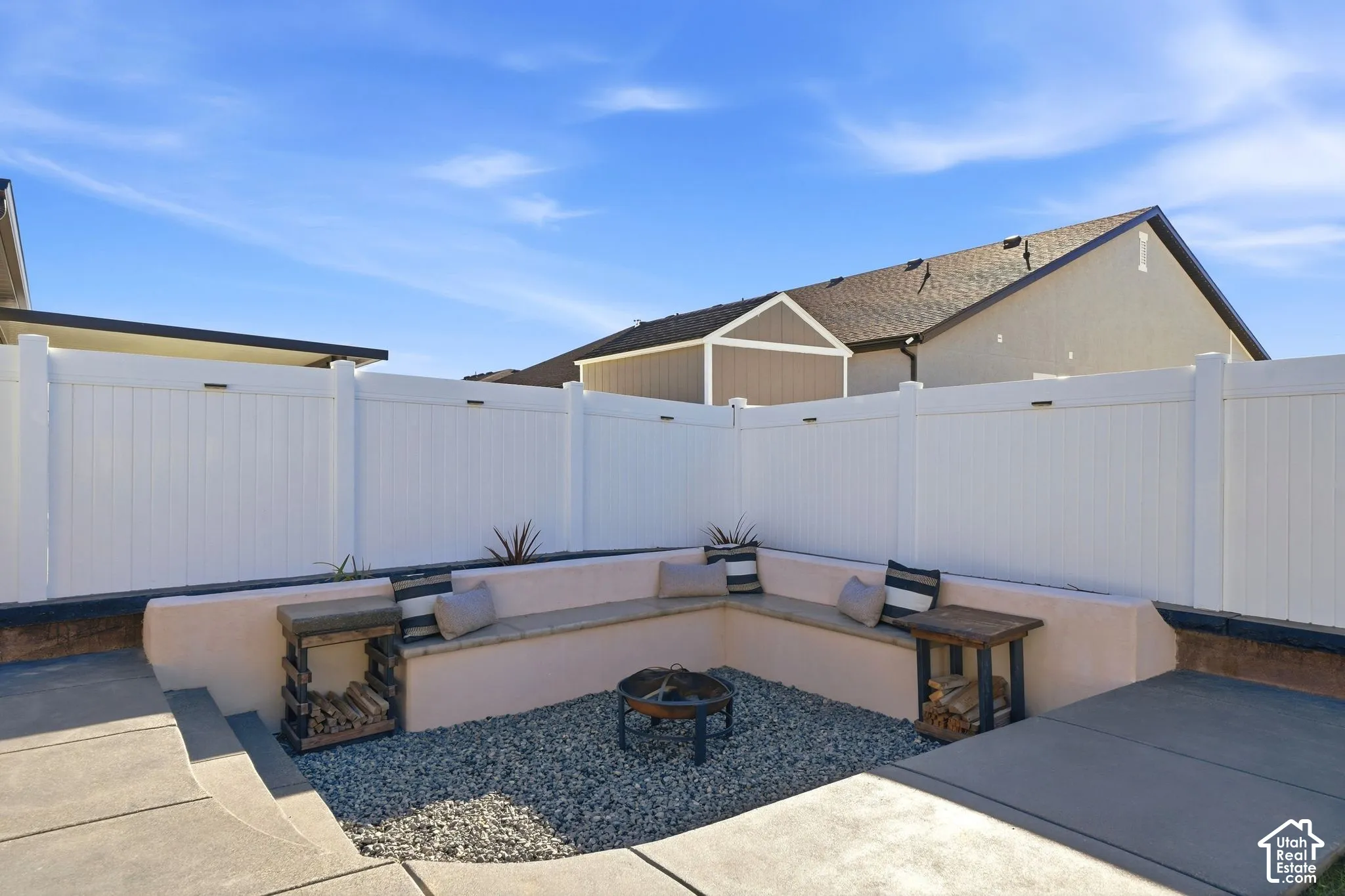 Entertaining backyard oasis featuring a curved built-in bench seating arrangement with cushioned comfort, gravel-filled fire pit area with metal surround, and white vinyl privacy fencing