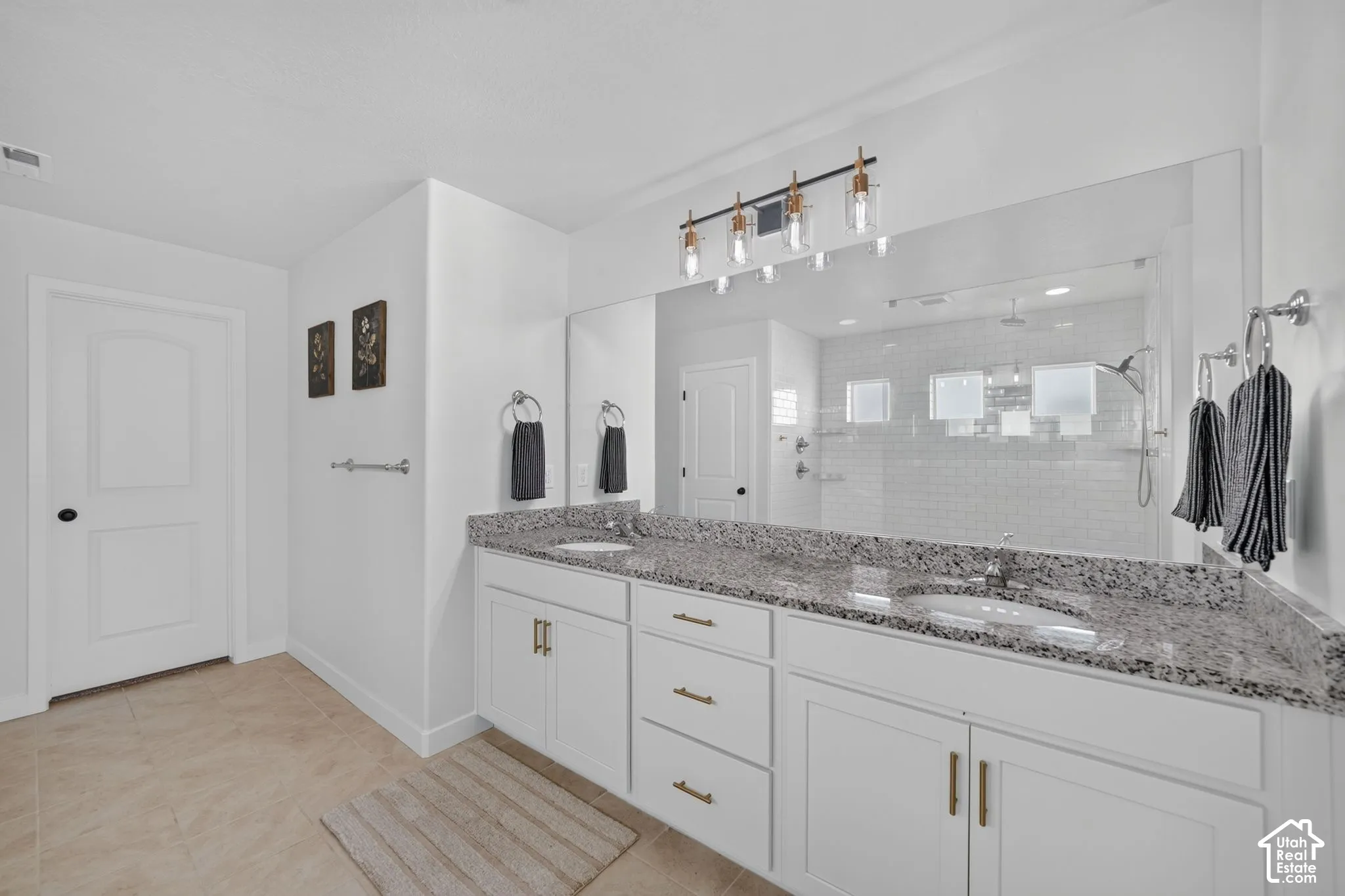 Dual vanity with granite countertop, white cabinetry with gold-tone hardware, a large wall-mounted mirror, and a walk-in shower with subway tile