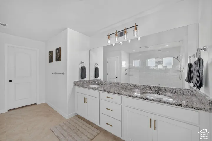 Dual vanity with granite countertop, white cabinetry with gold-tone hardware, a large wall-mounted mirror, and a walk-in shower with subway tile