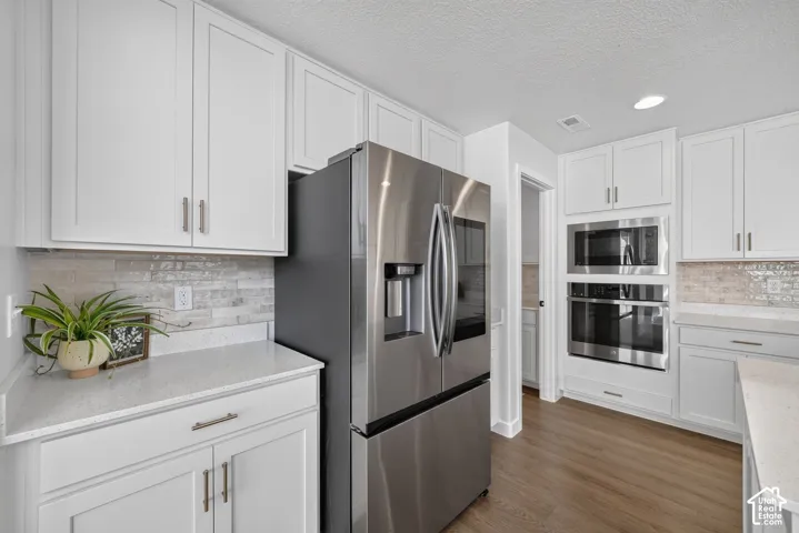 White shaker cabinetry, light-toned backsplash tiling, stainless steel appliances, light-toned countertops, and wood-finish flooring