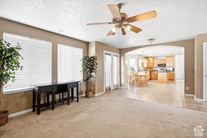 Carpeted living space with a ceiling fan, two windows with blinds, a double door, and an arched entryway leading to a kitchen with tile flooring