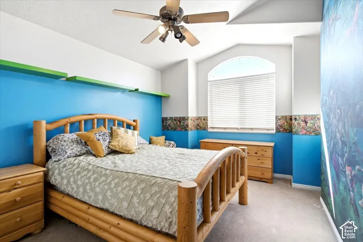 Arched window with blinds, vaulted ceiling, ceiling fan with integrated lighting, wall-mounted shelving, and carpeted flooring