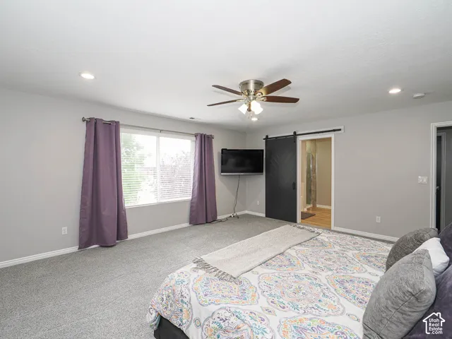 Humungous master bedroom featuring light gray walls, recessed lighting, a ceiling fan, a large window with blinds, and a black barn door leading to an adjacent bathroom area with wood-finish flooring.