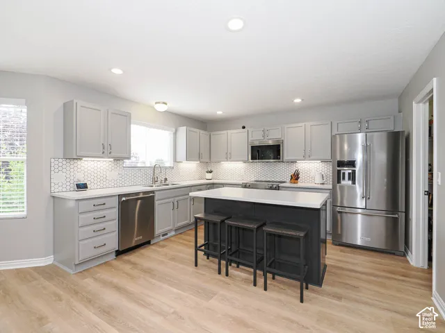 Modern kitchen featuring light gray cabinetry, new countertops, stainless steel appliances, a center island with seating, hexagonal tile backsplash, and light wood-finish flooring.