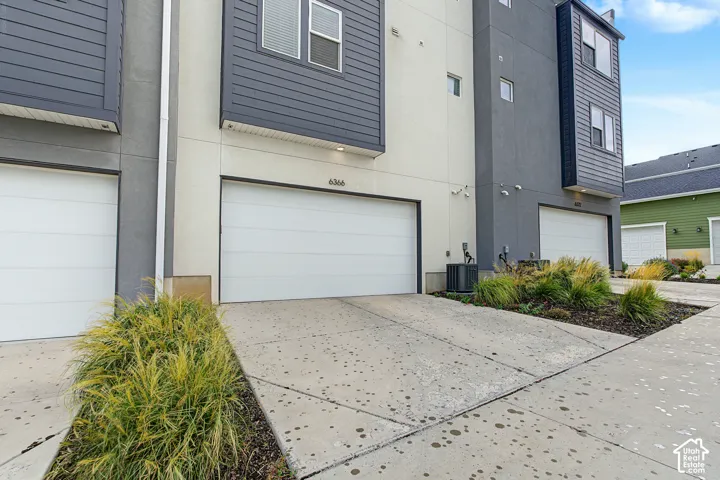 Modern driveway featuring a wide two-car garage with white panel doors, decorative concrete paving with aggregate finish, and contemporary architectural styling with mixed exterior cladding in charcoal and cream tones