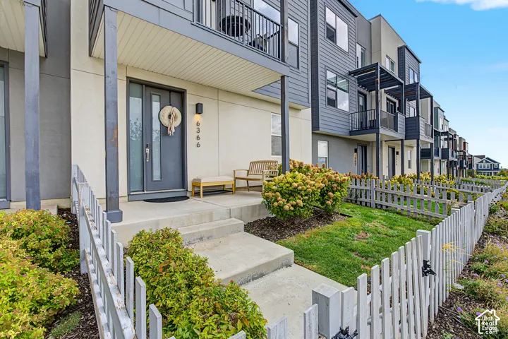 Modern townhome exterior with mixed material siding, concrete porch, dark gray entry door with vertical glass inserts, and a partial picket fence