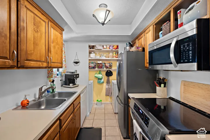 Galley kitchen featuring warm wood cabinetry, stainless steel appliances including a microwave and electric range, tile flooring, and ample counter space with a stainless steel sink