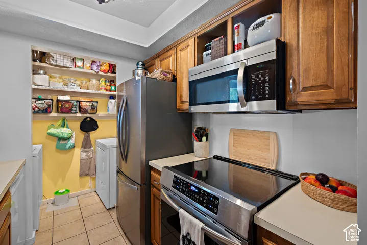 Stainless steel appliance suite, wood-finish cabinetry, light-toned countertops, tile flooring, and integrated pantry shelving
