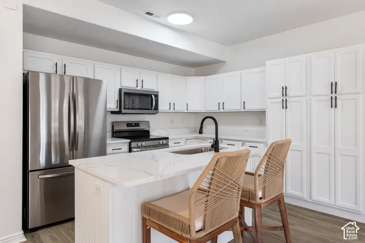 White cabinetry with black hardware, stainless steel appliances, a center island with a sink, white countertop surfaces, and wood-finish flooring