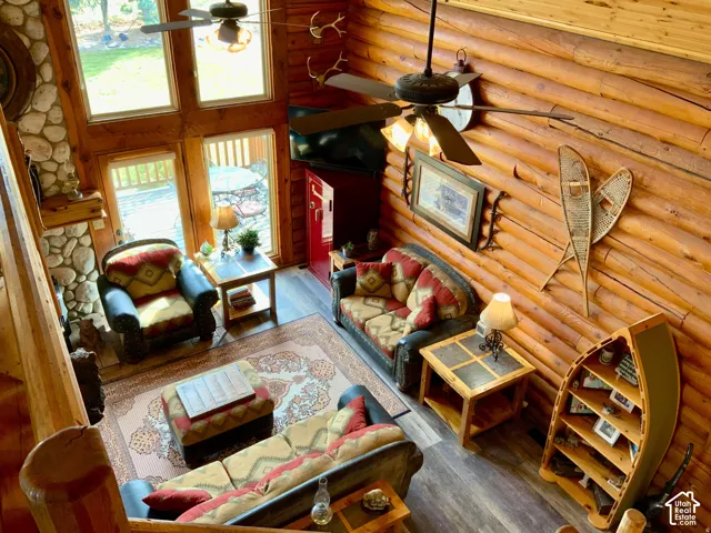 Vaulted living space featuring log walls, an exposed stone fireplace, wood-finish flooring, a ceiling fan, and large windows