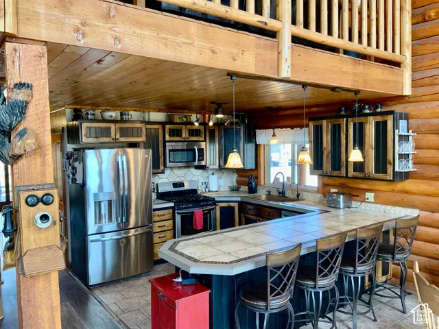 Log cabin interior featuring a kitchen with stainless steel appliances, a tiled countertop with an integrated breakfast bar, wood-paneled walls, and a wood ceiling with exposed beams